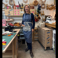 A smiling woman stands in a well-equipped artisan workshop wearing a blue apron over a dark long-sleeve top and jeans. The background is filled with tools, hanging woven baskets, storage drawers, and crafting supplies.