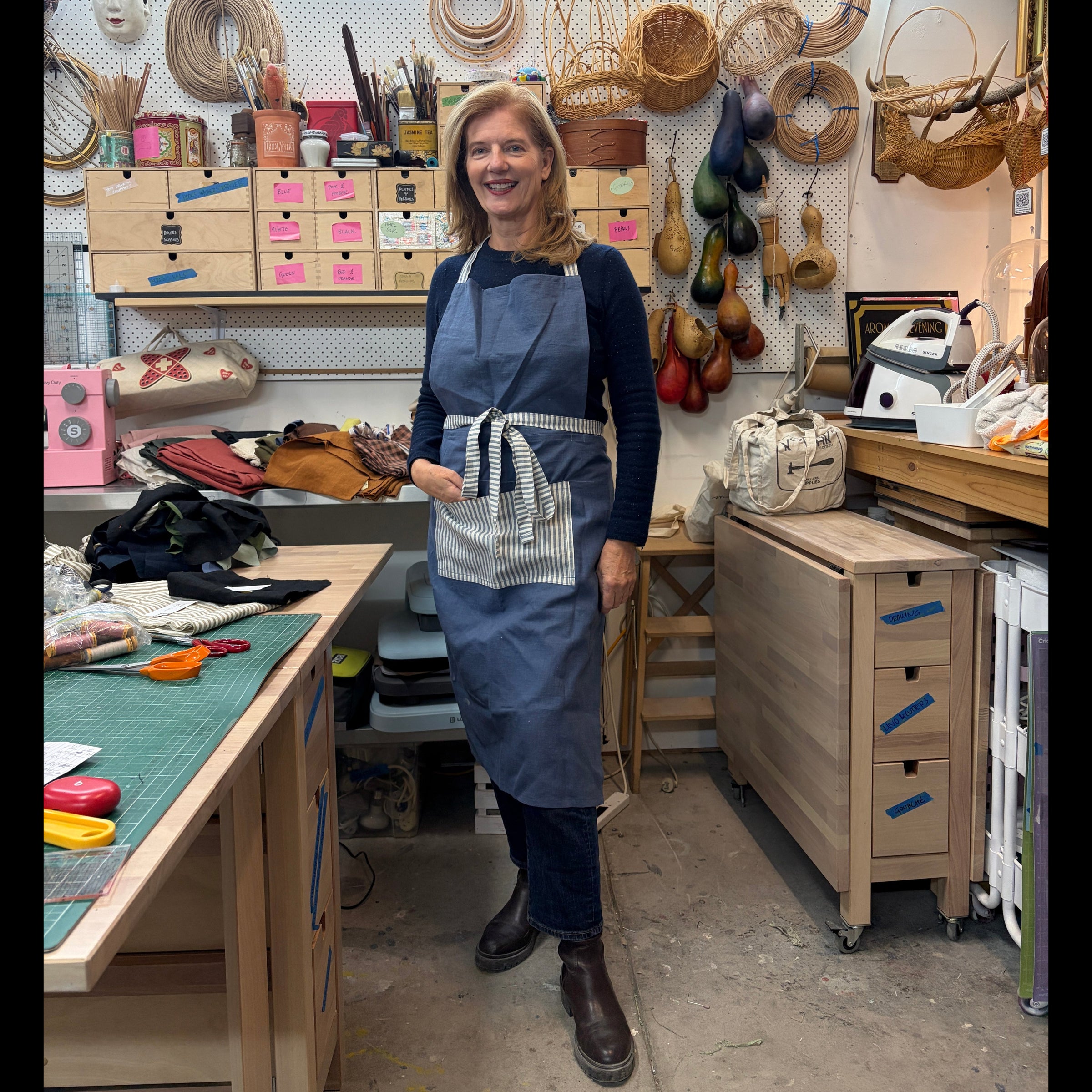 A smiling woman stands in a well-equipped artisan workshop wearing a blue apron over a dark long-sleeve top and jeans. The background is filled with tools, hanging woven baskets, storage drawers, and crafting supplies.