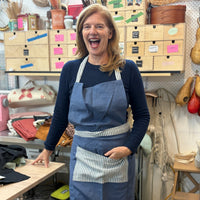 A woman smiling in an art studio wearing a blue apron with a striped pocket over a navy long-sleeve shirt. Shelves behind her hold wooden drawers, tools, and various materials.