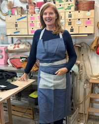 A smiling woman wearing a navy blue apron with a white and navy striped pocket over a navy long-sleeve shirt, standing in a craft or sewing workshop filled with wooden drawers, tools, and fabric.