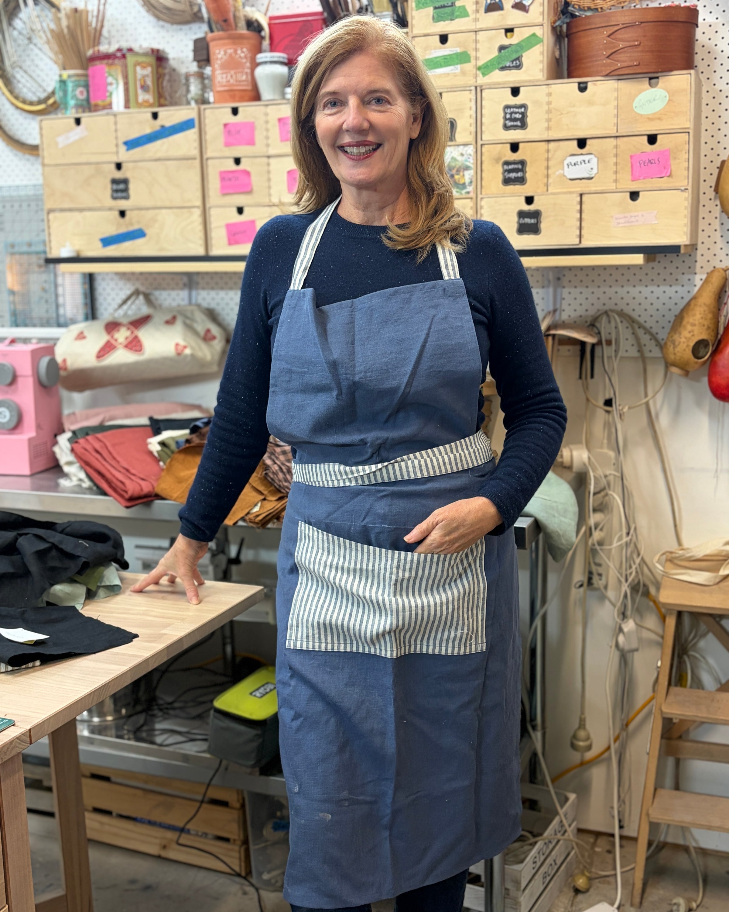 A smiling woman wearing a navy blue apron with a white and navy striped pocket over a navy long-sleeve shirt, standing in a craft or sewing workshop filled with wooden drawers, tools, and fabric.