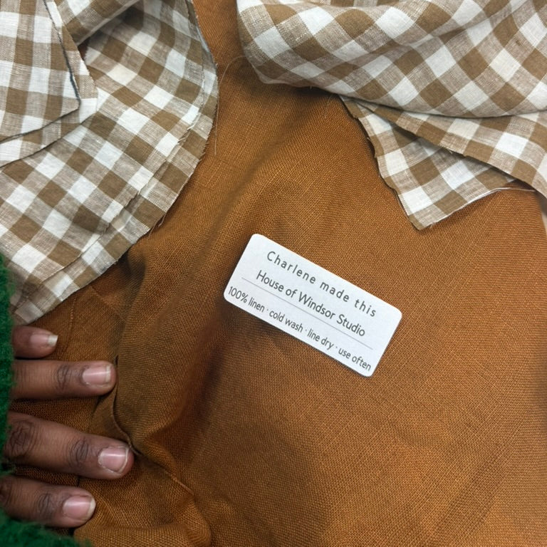 A close-up of a brown fabric garment or textile with a white label reading 'Charlene made this House of Windsor Studio 100% linen, cold-wash, line dry, use often.' A hand is visible holding the fabric, and a plaid brown-and-white cloth is draped across the top.
