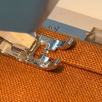 Close-up of a sewing machine presser foot stitching orange linen fabric.