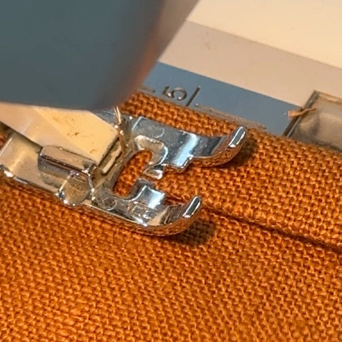 Close-up of a sewing machine presser foot stitching orange linen fabric.