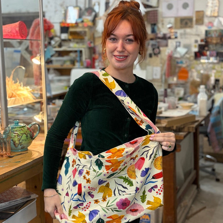A woman with red hair in a bun wearing a black velvet top and black skirt, holding a cream-colored shoulder bag with a colorful floral and botanical print in an art studio.