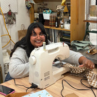 A woman with glasses and long dark hair sits behind a white sewing machine on a wooden table, sewing a piece of brown and white checkered fabric in a cluttered craft room with shelves and tools in the background.