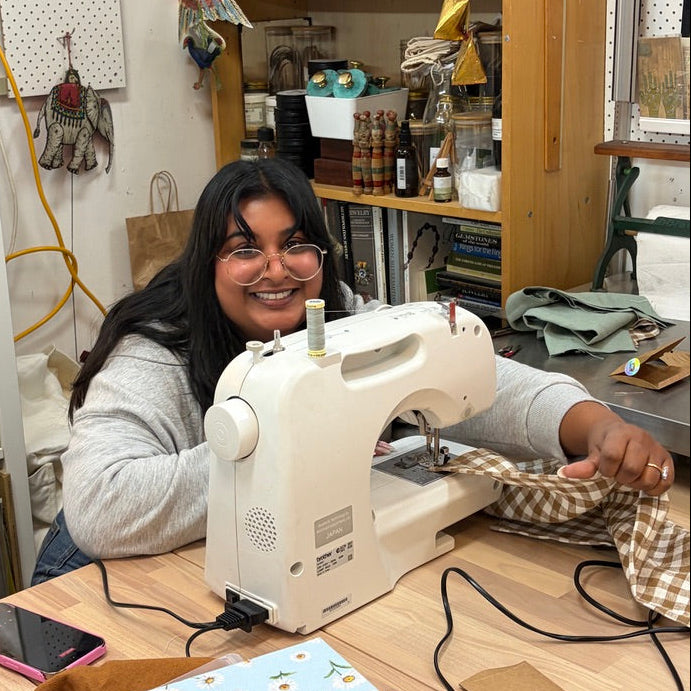 A woman with glasses and long dark hair sits behind a white sewing machine on a wooden table, sewing a piece of brown and white checkered fabric in a cluttered craft room with shelves and tools in the background.