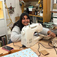 A woman with glasses and long dark hair sits behind a white sewing machine on a wooden table, sewing a piece of brown and white checkered fabric in a cluttered craft room with shelves and tools in the background.