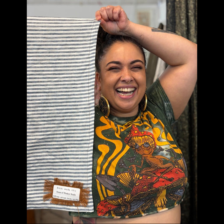 A woman smiling and holding up a striped linen tea towel next to her, showing a small woven label. The fabric has thin gray and white stripes, and the woman is wearing a graphic t-shirt with a colorful alien design.