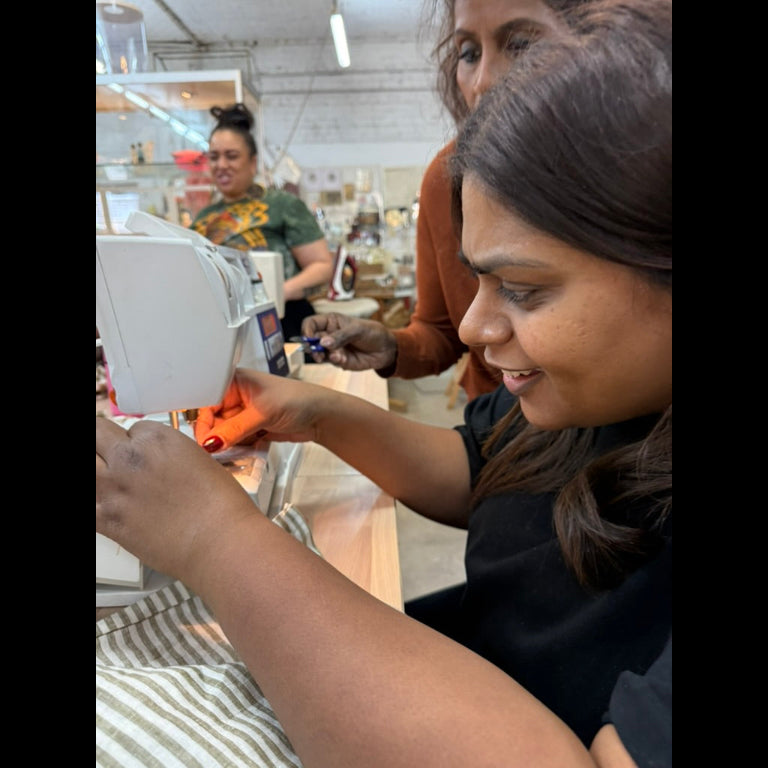 A woman in a black top uses a white sewing machine in a bright, active sewing workshop. Another person is visible in the background, and the scene is filled with the activity of the craft space.