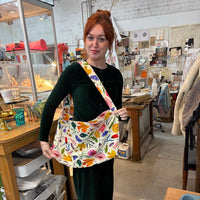 A woman with red hair stands in an art studio wearing a dark green velvet outfit and carrying a large canvas tote bag decorated with a colourful floral and bird pattern.