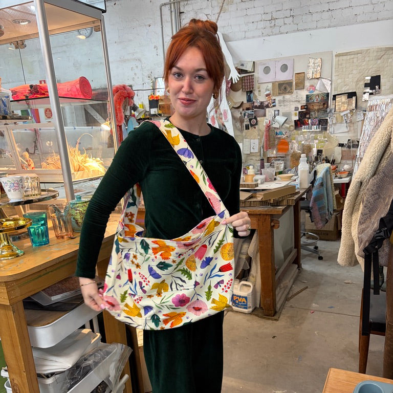 A woman with red hair stands in an art studio wearing a dark green velvet outfit and carrying a large canvas tote bag decorated with a colourful floral and bird pattern.