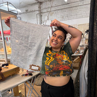 A woman in a workshop holds up a striped tea towel with black and white stripes. She is wearing a green graphic crop top and black pants, standing in front of an ironing table and other textile items.
