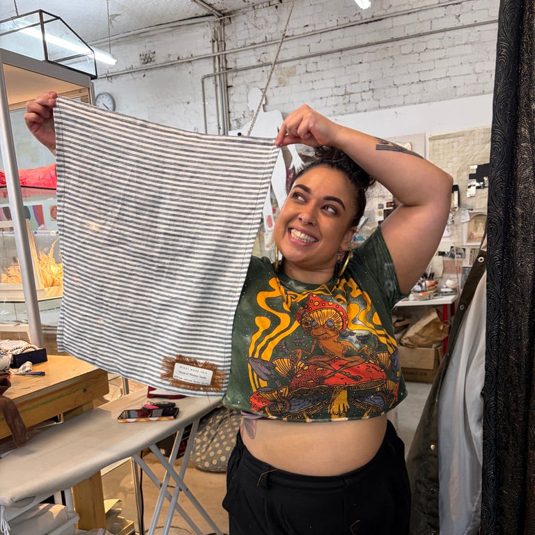 A woman in a workshop holds up a striped tea towel with black and white stripes. She is wearing a green graphic crop top and black pants, standing in front of an ironing table and other textile items.