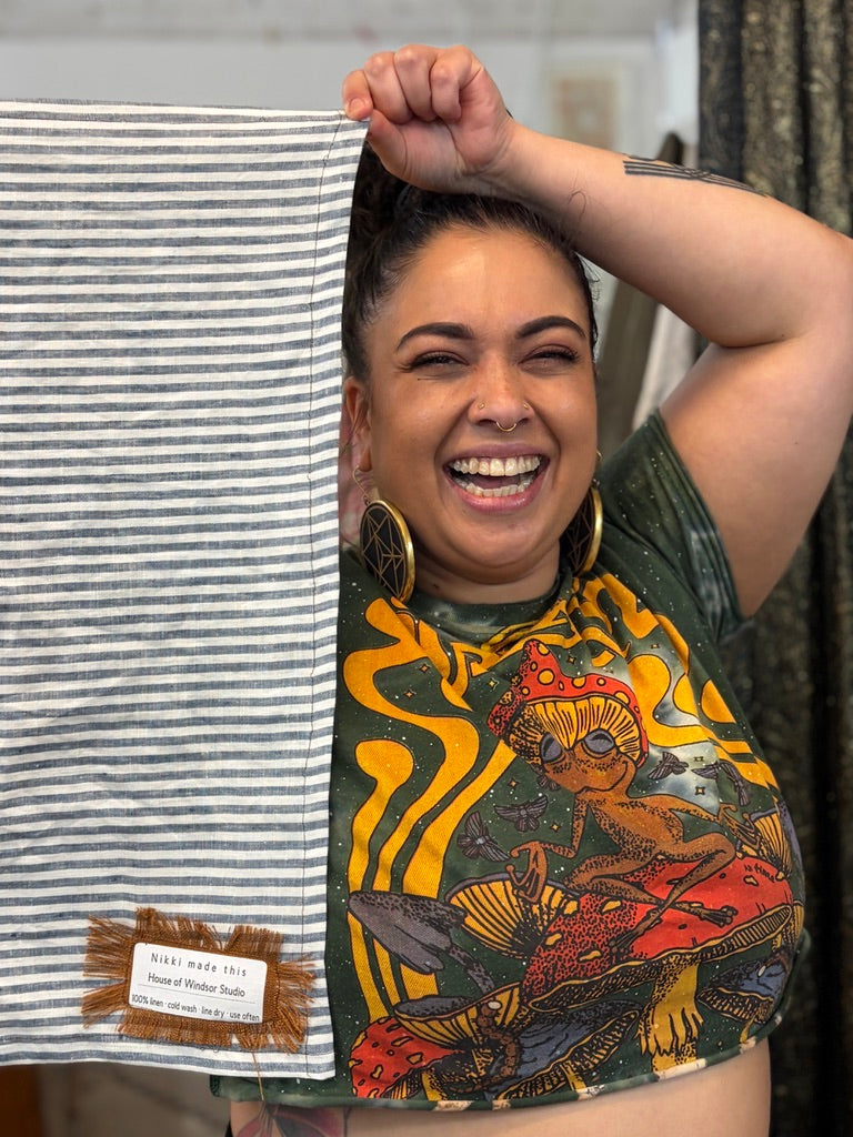 A woman smiling and holding up a striped fabric panel next to her, showing a small woven label. The fabric has thin gray and white stripes, and the woman is wearing a graphic t-shirt with a colorful alien design.