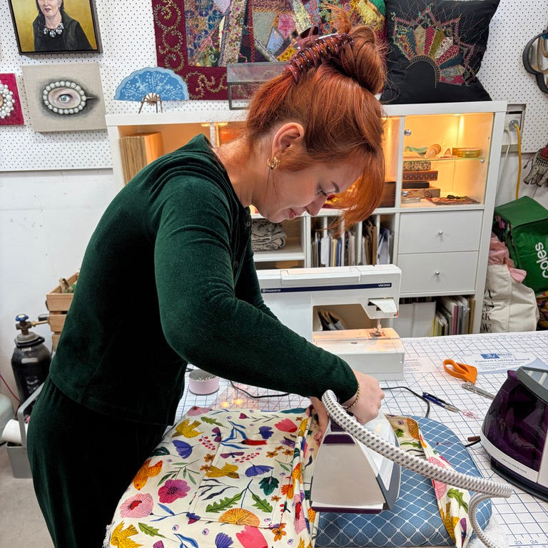 A woman with red hair tied up, wearing a dark green long-sleeve top, is ironing a colorful floral-patterned fabric bag on a wooden table in a sewing room.
