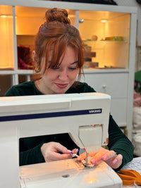 A woman with red hair in a bun is operating a white Viking sewing machine, sewing a small pink and white floral fabric piece. The background shows a white storage cabinet with warm lighting.