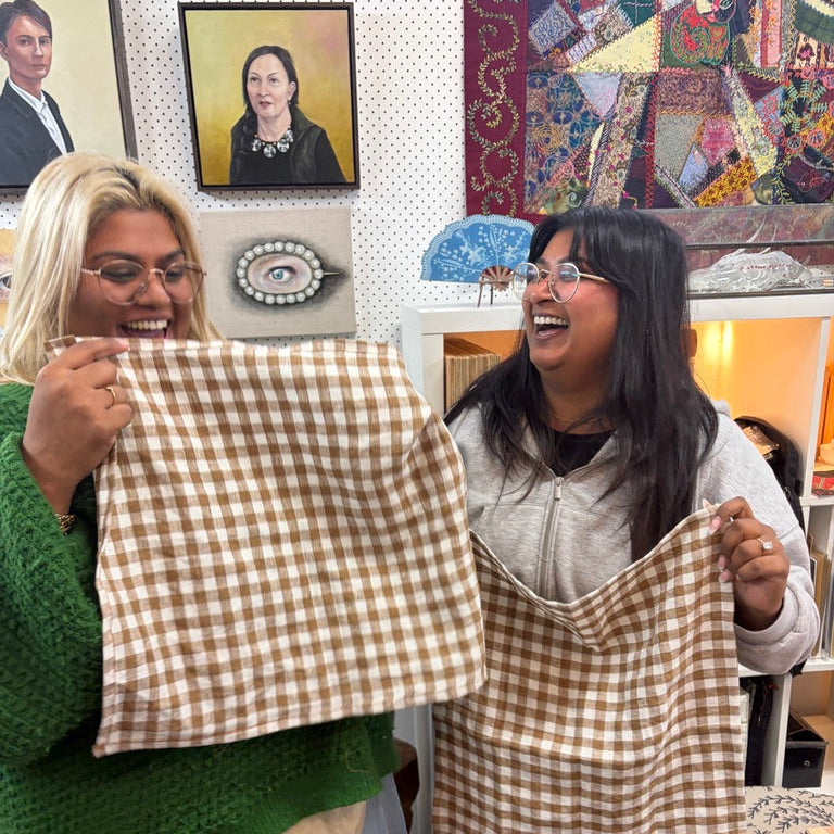 Two women laughing in a shop while holding up matching brown and beige gingham fabric cloths, with framed art and shelves in the background.