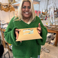A smiling woman with blonde hair and glasses holds up a sewing kit. She is wearing a chunky green sweater and light blue sweatpants in a creative workshop.