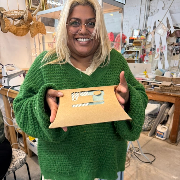 A smiling woman with blonde hair and glasses holds up a sewing kit. She is wearing a chunky green sweater and light blue sweatpants in a creative workshop.