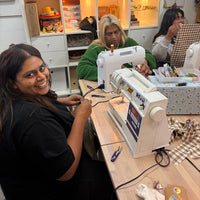 A woman in a black top smiles while working at a white sewing machine at a wooden table, with another woman seated behind her in a sewing room with shelves and quilts in the background.