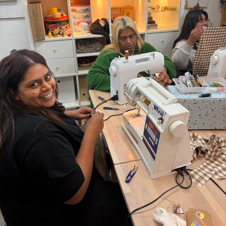 A woman in a black top smiles while working at a white sewing machine at a wooden table, with another woman seated behind her in a sewing room with shelves and quilts in the background.